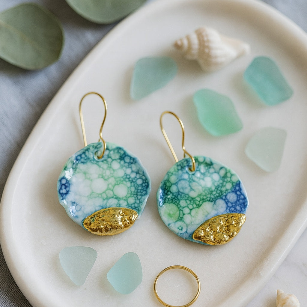 A pair of blue, green, and gold earrings on a white plate with sea glass and shells.