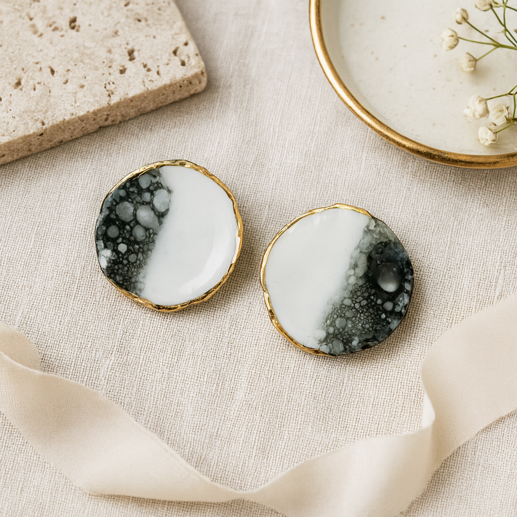 Pair of earrings with black, white, and gold design on a textured beige surface.